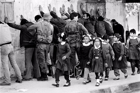 دانش‌آموزان مدرسه‌ای در حال عبور از کنار گروهی از مردان فلسطینی که توسط سربازان اسرائیلی در نوار غزه بازرسی بدنی می‌شوند، نوامبر ۱۹۸۶